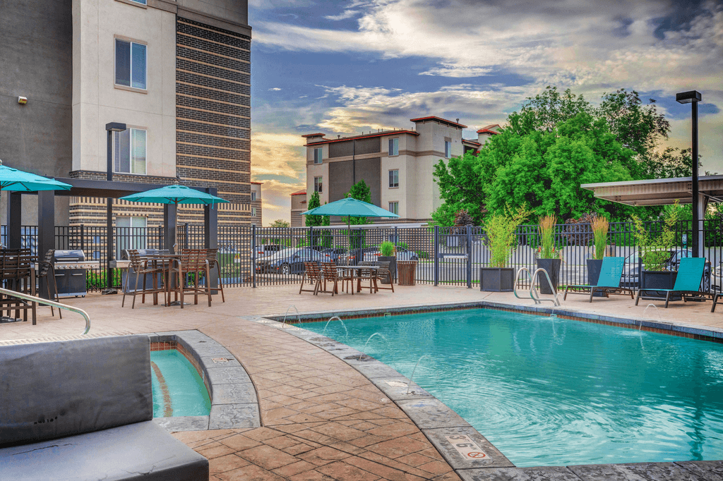 Swimming pool with tanning deck tables and chairs with blue umbrellas surrounded by building exterior and native landscape.
