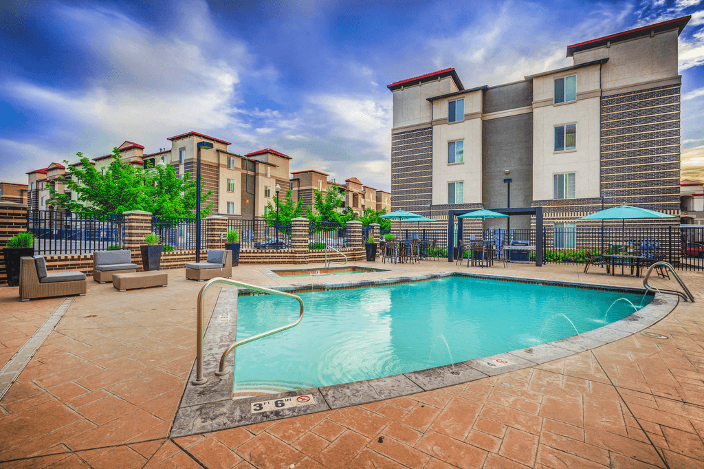 Swimming pool with tanning deck tables and chairs with blue umbrellas surrounded by building exterior and native landscape.