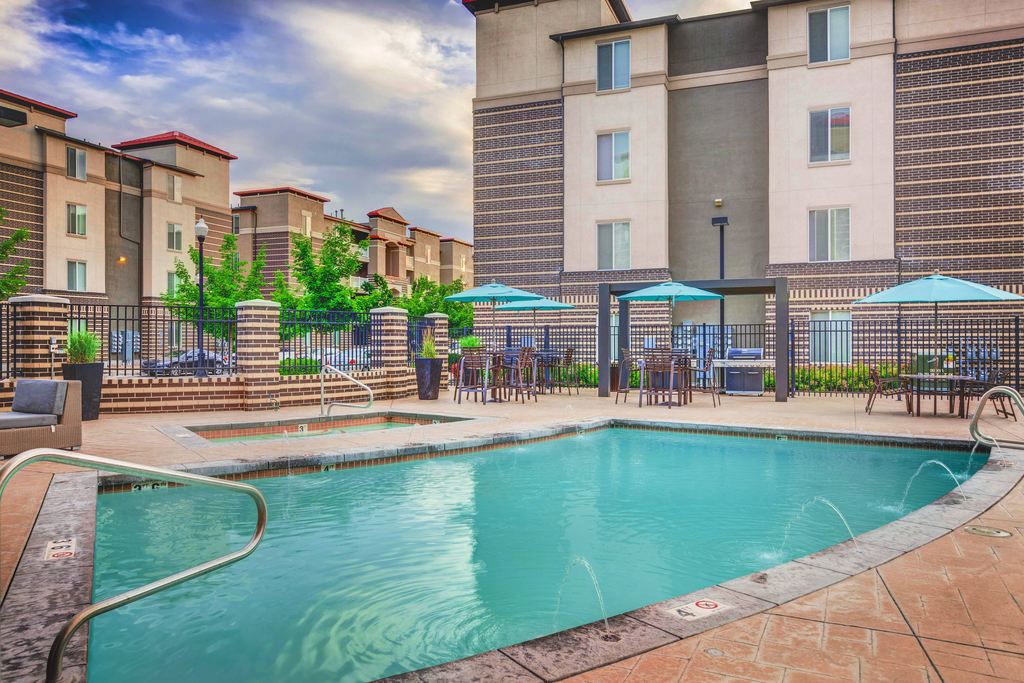Swimming pool with tanning deck, tables and chairs with blue umbrellas surrounded by native landscape and building exterior