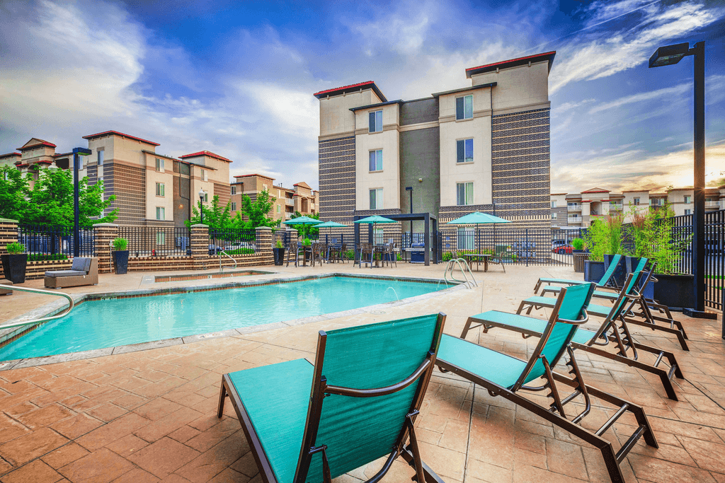 Swimming pool with tanning deck tables and chairs with blue umbrellas, blue lounge chairs and surrounded by building exterior and native landscape.