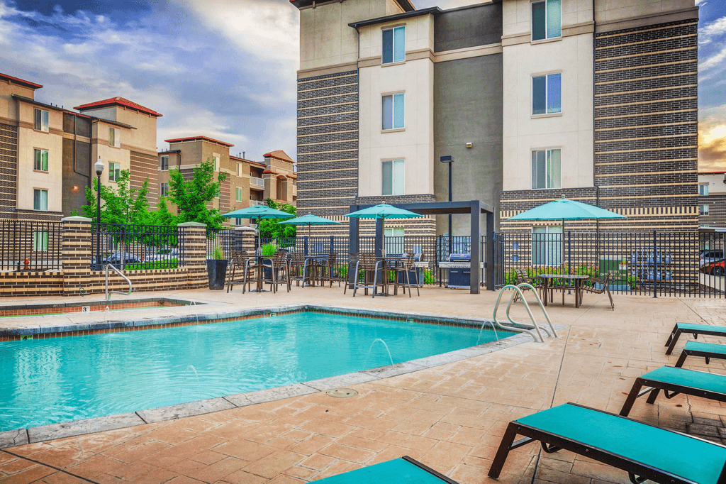 Swimming pool with tanning deck tables and chairs with blue umbrellas surrounded by building exterior and native landscape.