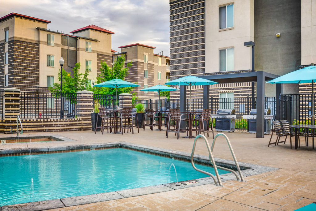 Swimming pool with tanning deck, tables and chairs with blue umbrellas surrounded by native landscape and building exterior