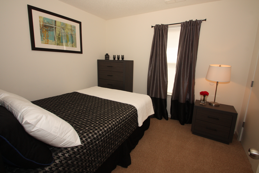 The furnished bedroom is carpeted with a queen bed, dresser and nightstand. The bed has a houndstooth comforter with white throw blanket, and a white pillow. There are black curtains hanging on the window and framed artwork on the wall.