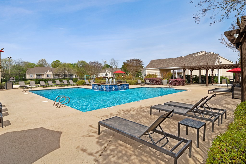 Community pool with sundeck, lounge chairs, surrounded by black metal fence with trees, and building exteriors in the background