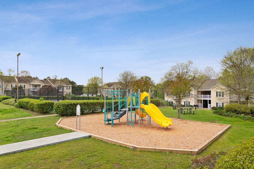 Playground in bed of mulch and yellow slide with a sports court in the back