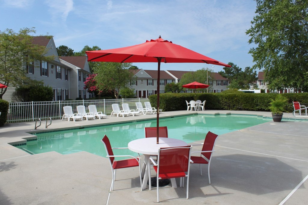 Community swimming pool with four red chairs and a red umbrella surrounding a table