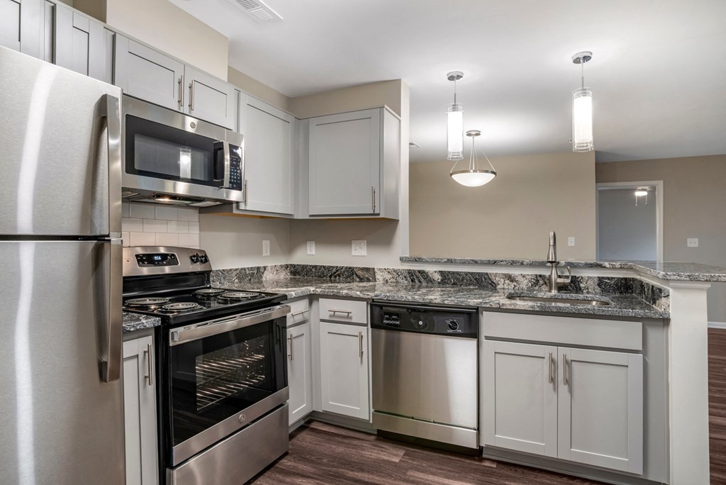 A vacant, renovated kitchen with gray walls, hardwood style flooring, ash gray cabinets with brushed nickel pulls, granite countertops, a single undermount sink with gooseneck faucet, stainless steel appliances, white subway tile backsplash, and a breakfast bar that opens to the dining room. Appliances include refrigerator, built-in microwave, stove/oven combo, and a dishwasher.