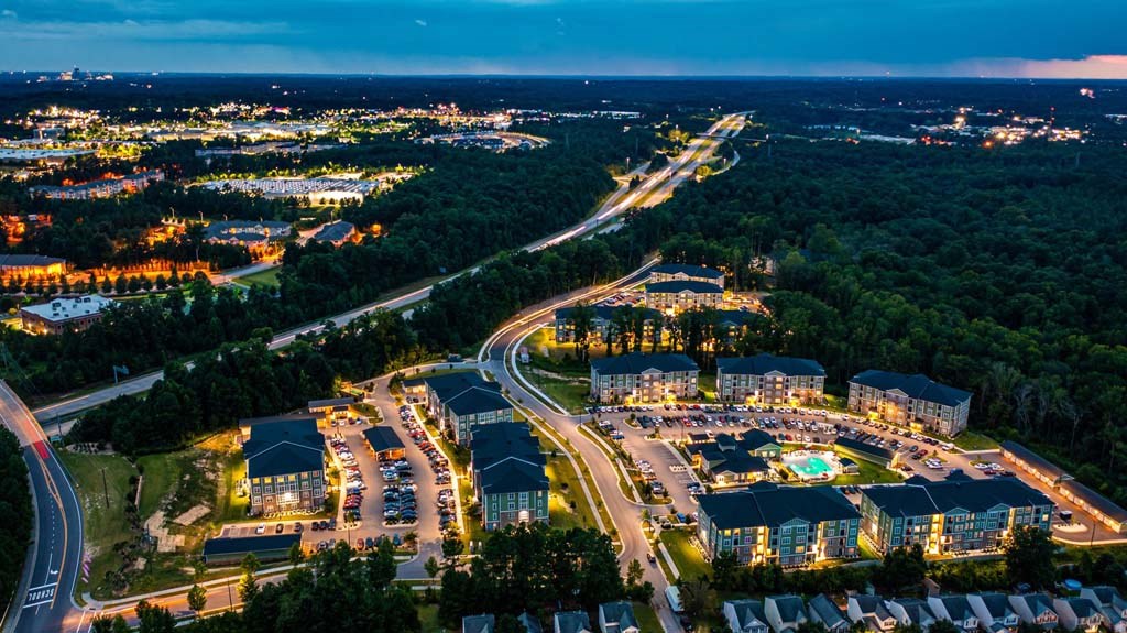 A nighttime aerial view of a resort with a lit swimming pool and cars parked in the parking lot at Foxwood Apartments, Raleigh-Durham, NC, 27616