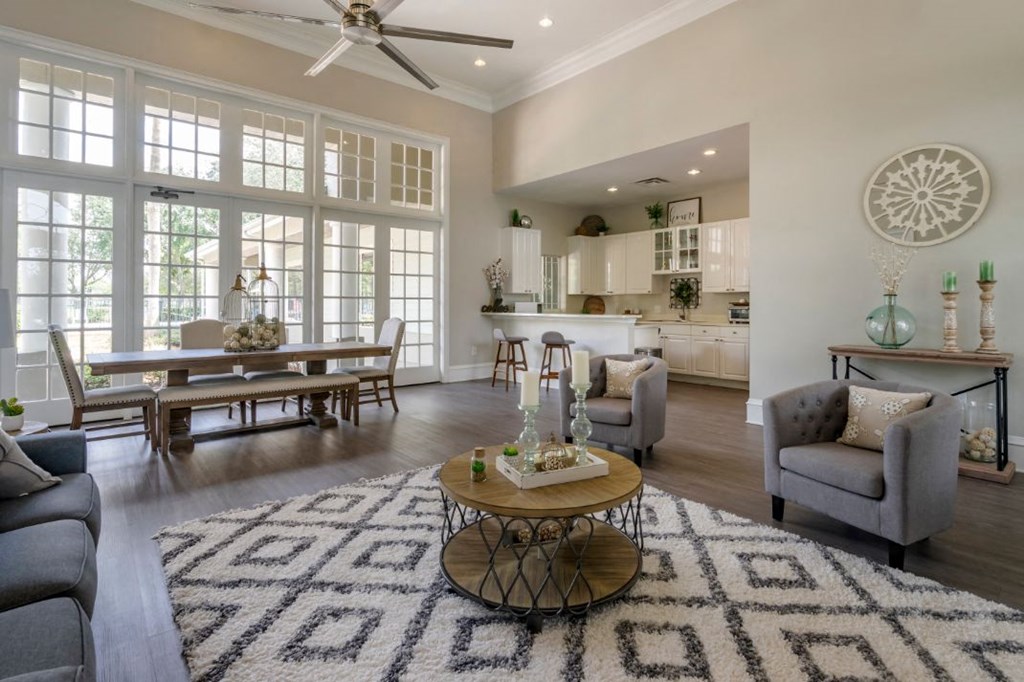 Clubhouse lounge with couch, chairs, coffee table, rug, picnic table, multi speed ceiling fan, large windows for natural lighting, and clubhouse kitchen in the background