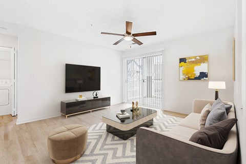 A living room with a brown ottoman, a grey couch, a black and white rug, a wooden floor, a flat screen TV, a ceiling fan, and a painting on the wall.
