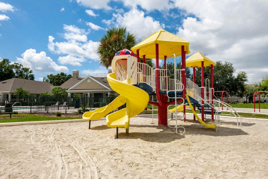 Blue Red and Yellow Playground on Sand with Building Exterior and Treeline in the Background