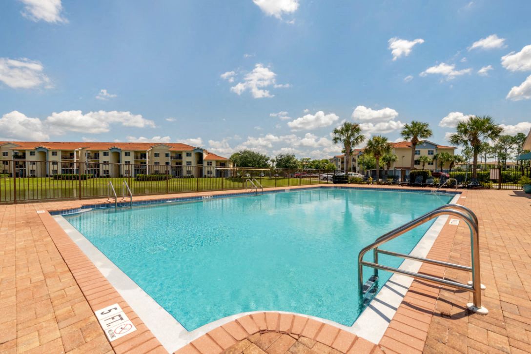 Fenced in Swimming Pool and Sun Deck with Lounge Chairs with Palm Trees and Building Exteriors in the Background