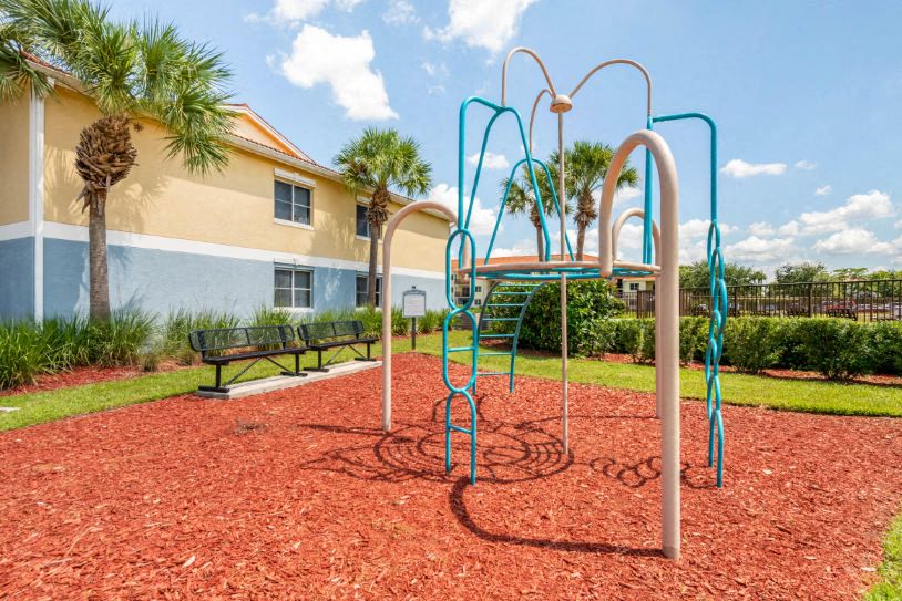 Playground with Green Accents on Mulch with Building Exterior and Benches with Treeline in the Background
