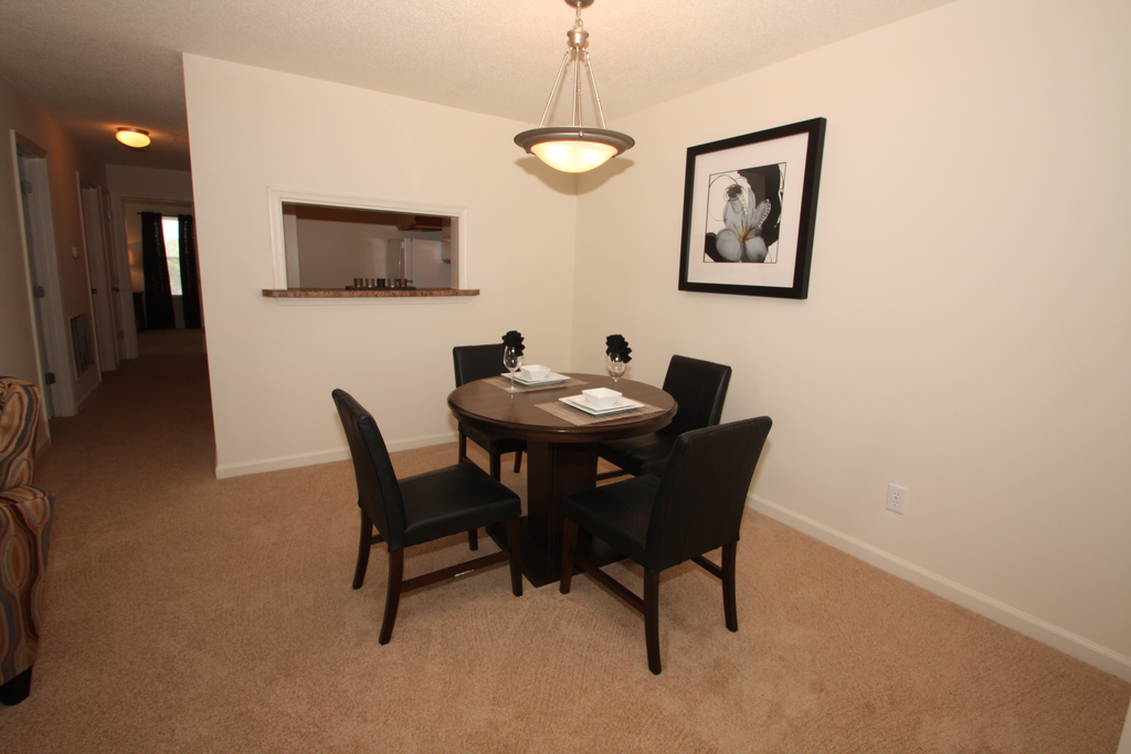 The furnished dining room has carpet throughout. There is a round wooden table with four black chairs positioned around it. There is a place setting for two on top of the table. There is a small breakfast bar that opens to the kitchen.