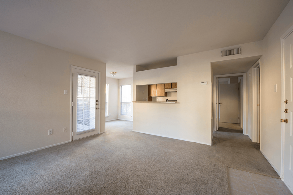 the living room and kitchen of an apartment with carpeting and white walls