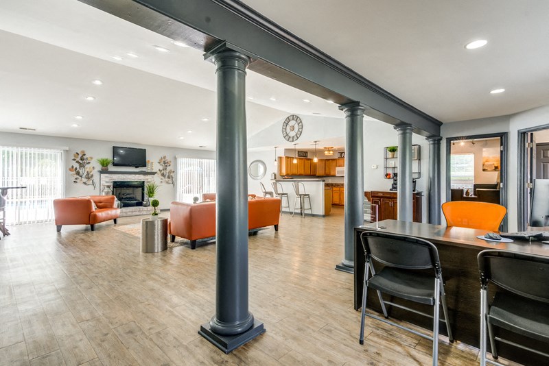 The interior of the Clubhouse/Leasing Office has hardwood style flooring, light gray walls with dark gray columns and trim. It is furnished with an orange couch, matching chair, area rug, a leasing desk with an orange chair and two black folding chairs. A kitchen area is located in the background.