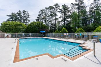 The swimming pool is rectangular in shape surrounded by an expansive sundeck with gray and blue lounge chairs positioned throughout. There is a blue umbrella in the corner of the pool area. The entire pool area is fenced in and is surrounded by grass, trees, and bushes.
