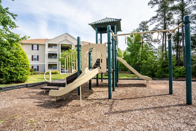 The community playground is surrounded by a large area of mulch, grass and trees. The play structure is green and tan with several climbing features, stairs and two single slides. An apartment building is located in t he background.