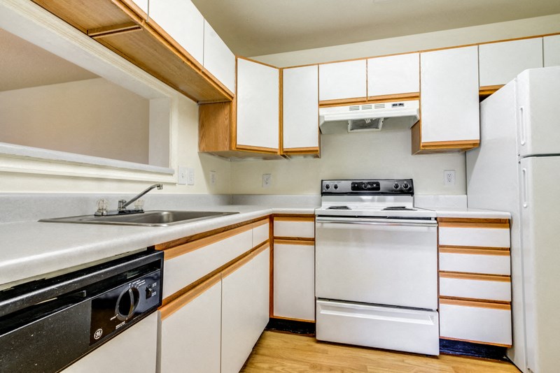A vacant kitchen with hardwood style flooring, white walls, white cabinets with light wood trim, white appliances, light countertops, a single basin sink and a breakfast bar area located behind the sink. Appliances include a refrigerator, a stove/oven combo and a dishwasher.