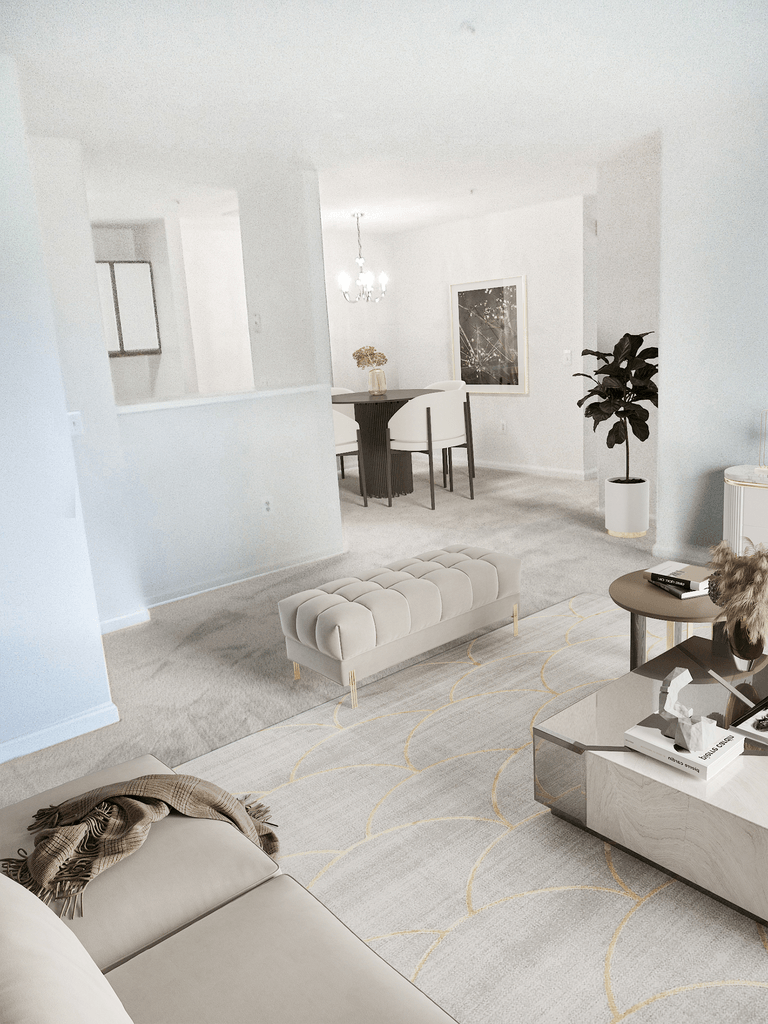 Vacant living room with tan carpet, white walls, and ceilings. The kitchen is in the background with a chandelier hanging. Virtually staged with a couch, ottoman, accent rug with a coffee table, standing potted plant, and in the dining room a table with chairs and art hanging on the wall.