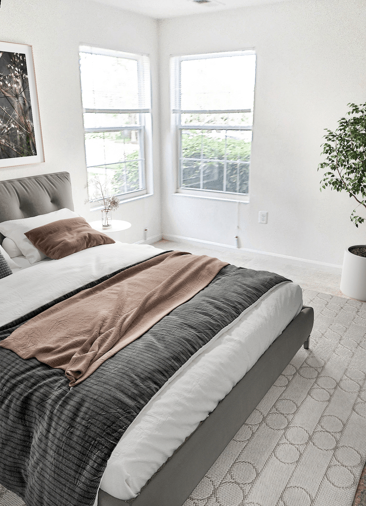 Bedroom with a bed and comforters next to two windows and a plant