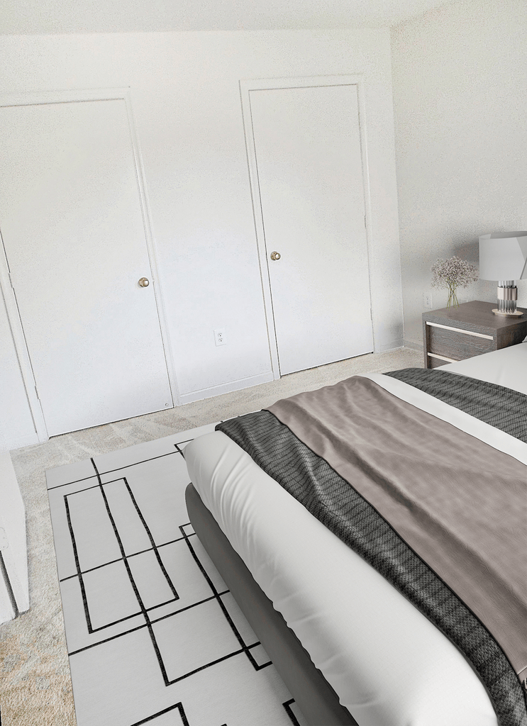 Vacant bedroom with tan carpet, white walls, and ceilings. Two doors are in the back of the room. Virtually staged wit a bed, side table with lamp, and an accent rug.