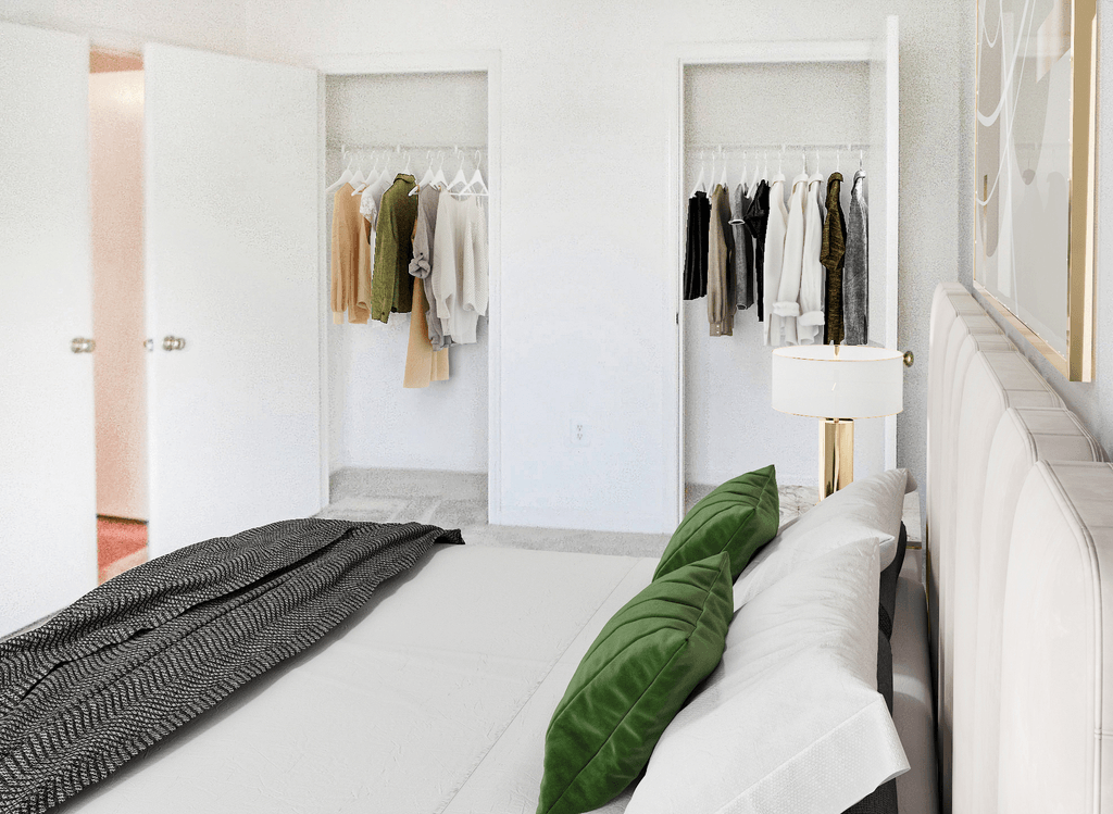 Vacant bedroom with tan carpet, white walls and ceiling. Double closets rest in background. Virtually staged with a bed, side table with a lamp, wall art, and clothes hanging in the closet.