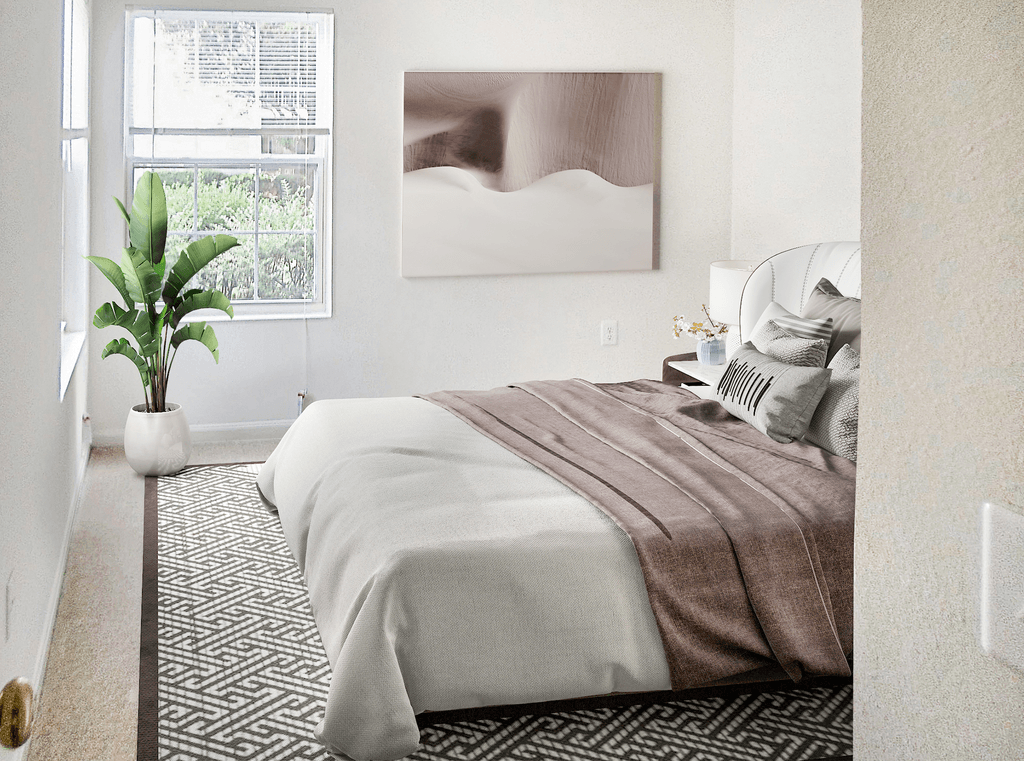 Vacant apartment bedroom with light tan carpet, white walls, and ceilings. The room has two windows on opposite walls. Virtually staged with a bed, end table with a lamp and flower, a potted standing plant in the corner and wall art next to the window.