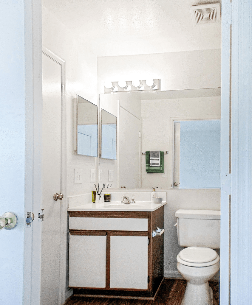 Vacant bathroom with vanity, sink, and toilet. Linoleum floor throughout with a large mirror on rear wall. Virtually staged with hand towels and soap on the countertop.