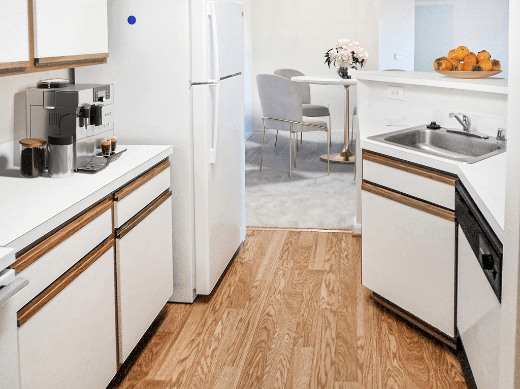 Vacant apartment kitchen with hardwood-style flooring, white wood trimmed cabinets, white appliances and countertops. Virtually staged with a coffee machine, a bowl of oranges on the bar top and a dining table with four chairs in the background.