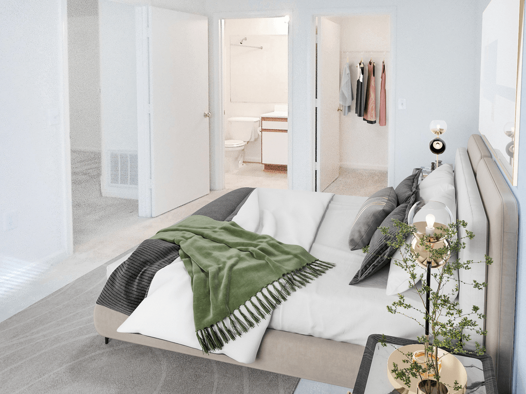 Vacant bedroom with tan carpet and white walls throughout. The closet and en-suite bathroom are in the background of the room. Virtually staged with a bed, wall art, accent rug, side table with lamp, and clothes hanging in the closet.