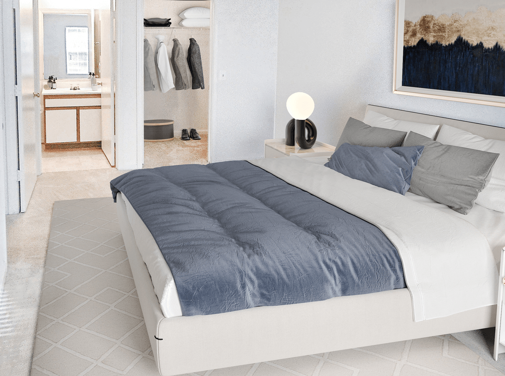 Vacant bedroom with tan carpet and white walls throughout. The closet and en-suite bathroom are in the background of the room. Virtually staged with a bed, wall art, accent rug, side table with lamp, and clothes hanging in the closet.