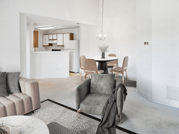 Vacant living room with tan carpet, white walls, and ceilings. The kitchen is in the background with a chandelier hanging over the dining area. Virtually staged with a couch, decorative chair, accent rug and coffee table, with dining table and four chairs in the background under the chandelier.