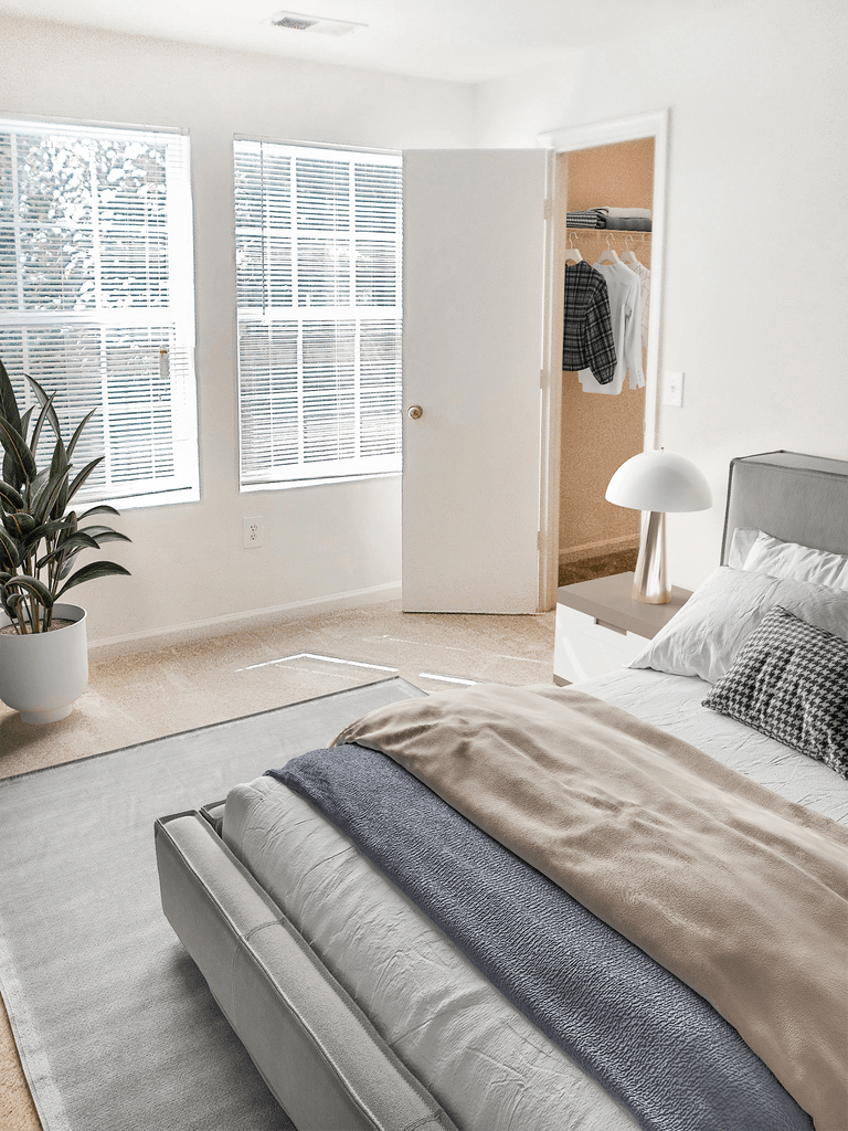 Vacant apartment bedroom with light tan carpet, white walls, and ceilings. The room has two windows on the back wall. Virtually staged with a bed, side table with a lamp, accent rug, potted standing plant and clothes hanging in the closet.