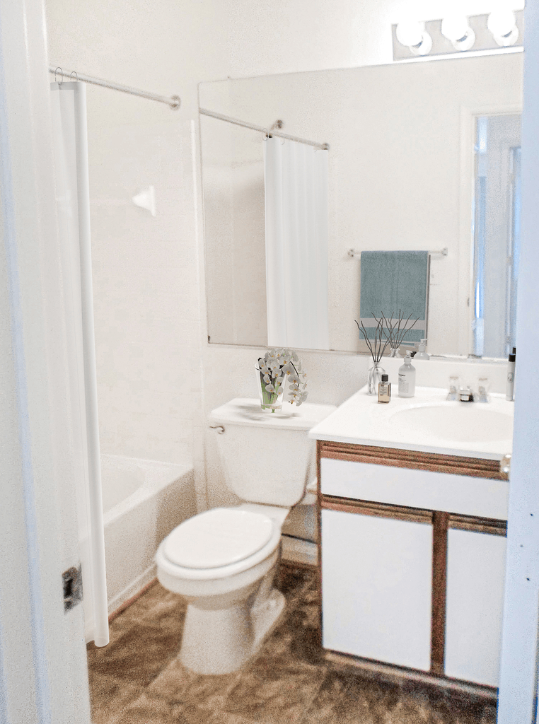 Bathroom with white countertops and sink with white cabined with wood trim, bathtub, toilet and large vanity mirror virtually staged with a white shower crutain, a blue towel on the towel bar, flowers on the toilet base and soap on the countertop
