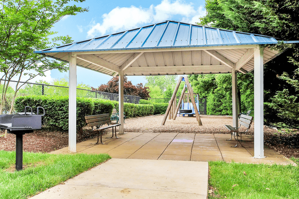 Outdoor pavilion with concrete floors and two benches next to a grill with playground in the background