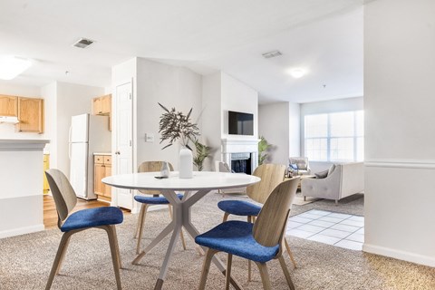 Vacant staged living room with wall-to-wall carpet. Door that lead to patio, vaulted ceiling and fireplace. View of kitchen in the background with white appliances and brown cabinets. Dinning table and couches set up in the back with mounted tv above the fireplace.