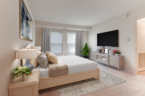 A bedroom with a large bed, a nightstand with a lamp, a dresser with a mirror, and a TV mounted on the wall.