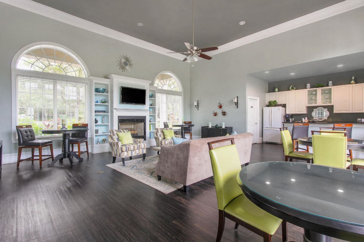 Clubhouse Seating Area with Large Windows on either side of Fireplace with TV and Shelves Surrounding Fireplace Multi Colored Accent Chairs Grey Couch and Dining Room Tables with Green Chairs and Clubhouse Kitchen in the Background