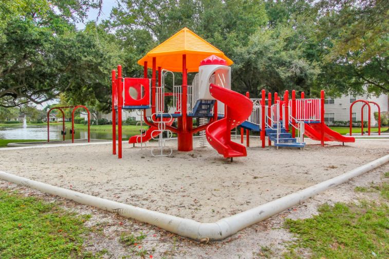 Blue Red and Yellow Playground on Sand with Building Exterior and Treeline in the Background