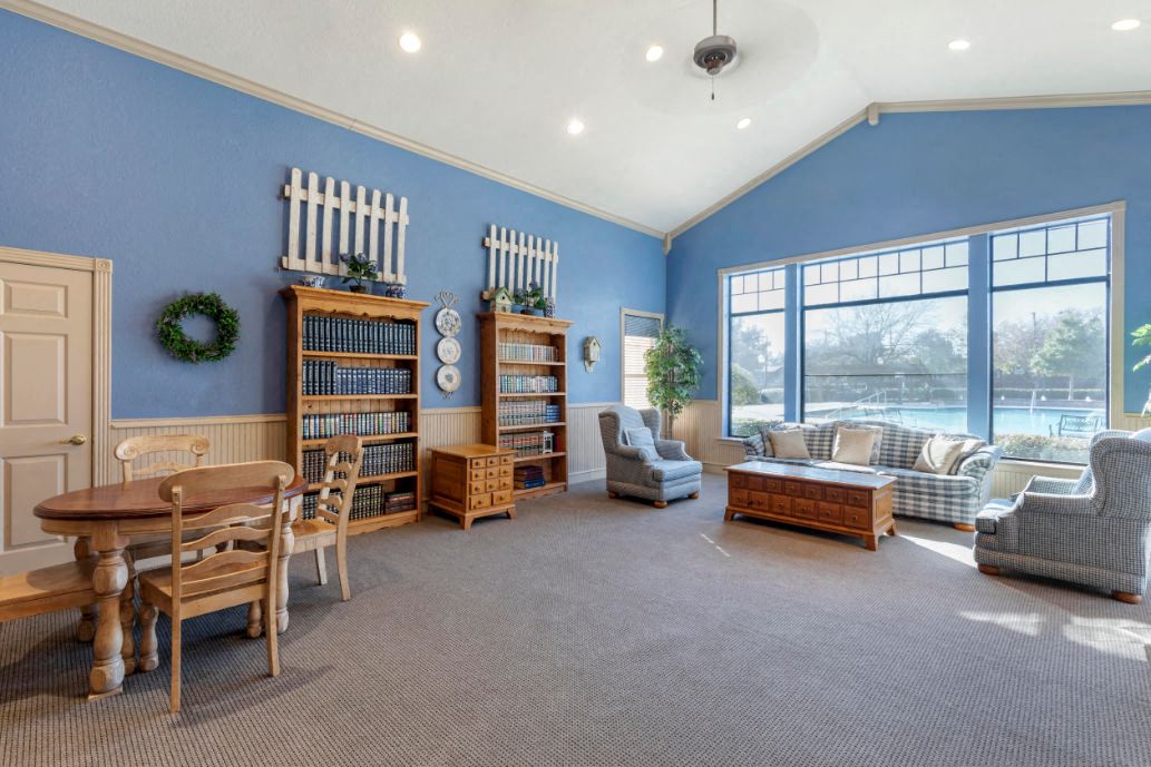 A living room with a dining table and chairs, a bookcase, and a window with a view of trees.