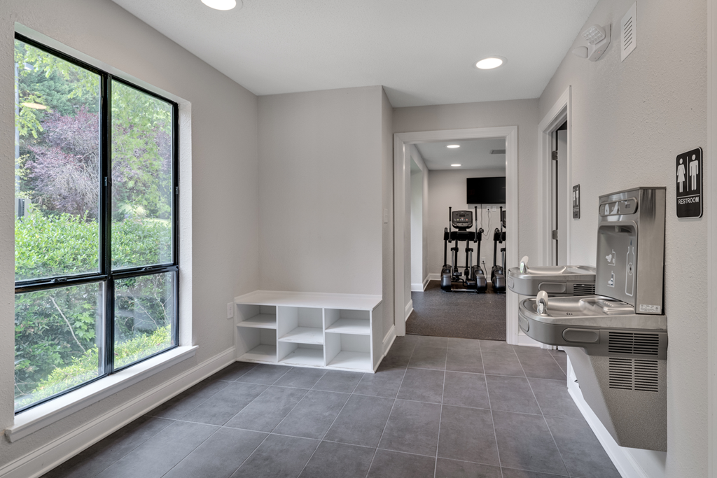 A room with a window, a bench, and water fountains outside of the fitness center.