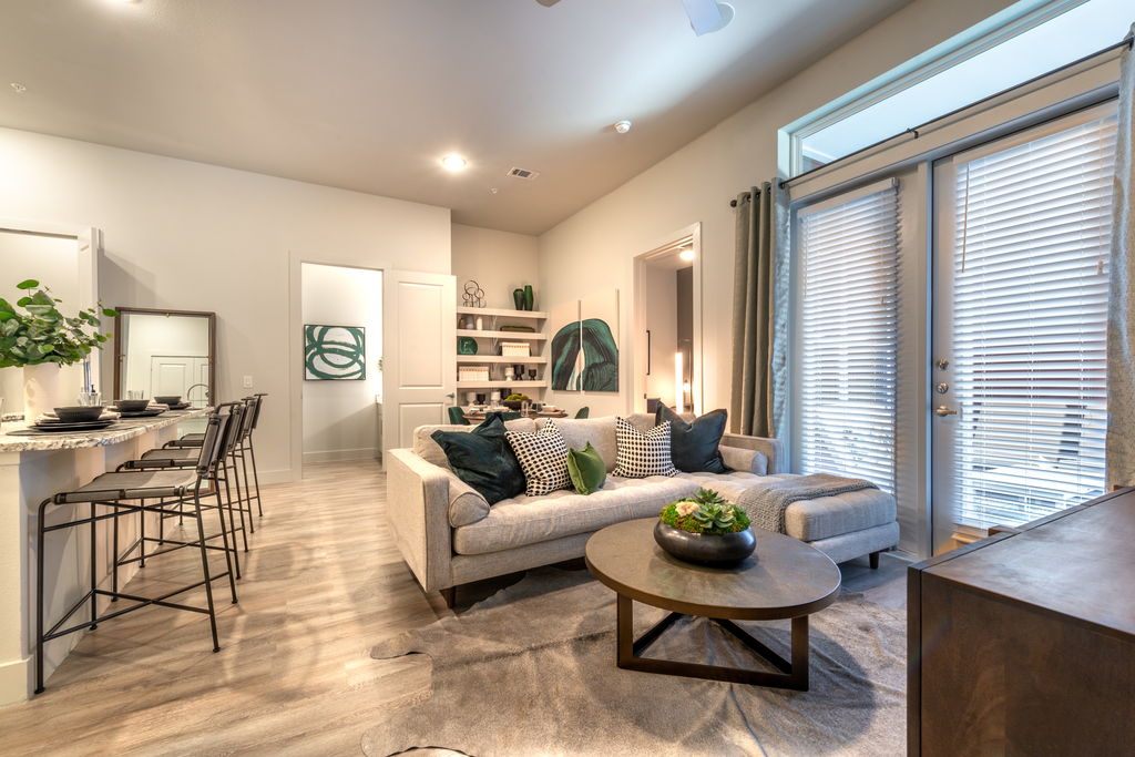 Staged livingroom with beige L-shaped couch, coffee table, accent rug, wood style flooring, bar stools are breakfast bar and built in book shelves in background