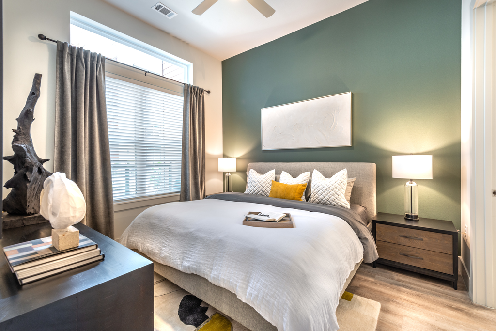 Staged bedroom with bed, accent rug, wood style flooring, night stands with lamps, dark dresser, accent wall, large window with blinds and curtains