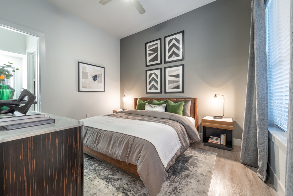 Staged bedroom with bed, accent rug, wood style flooring, side tables with lamps, and gray accent wall with art