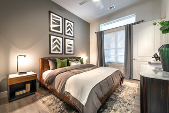 Staged bedroom with bed, accent rug, wood style flooring nightstands with lamps, gray accent wall with art, ceiling fan and large window with blinds and curtain - Photo Gallery 41