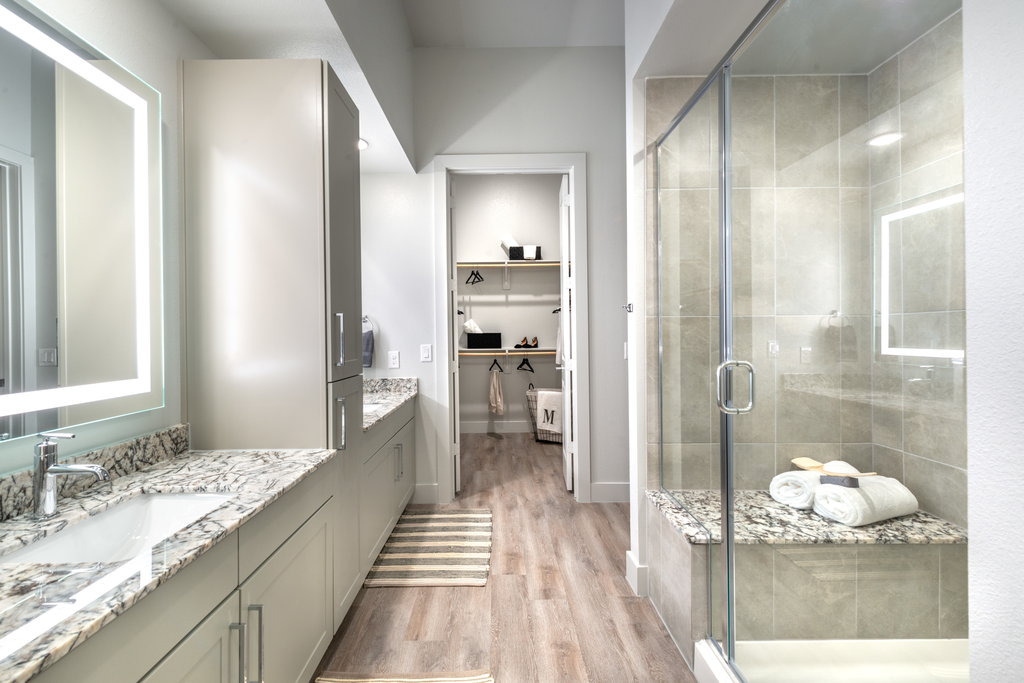 Staged bathroom with dual vanities, granite countertops, taupe cabinetry, walk in shower with bench, wood style flooring and direct access to walk in closet