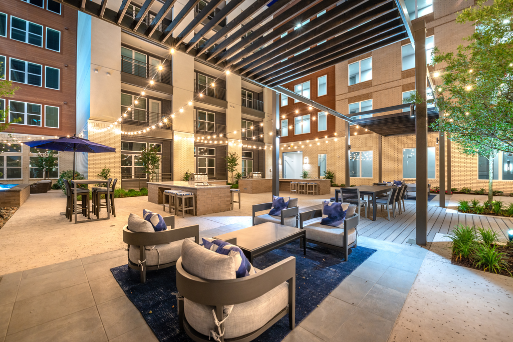 Outdoor grill area and lounge with gas grills, pergola seating, high top tables with umbrella and hanging lights with surrounding building exterior and native landscape