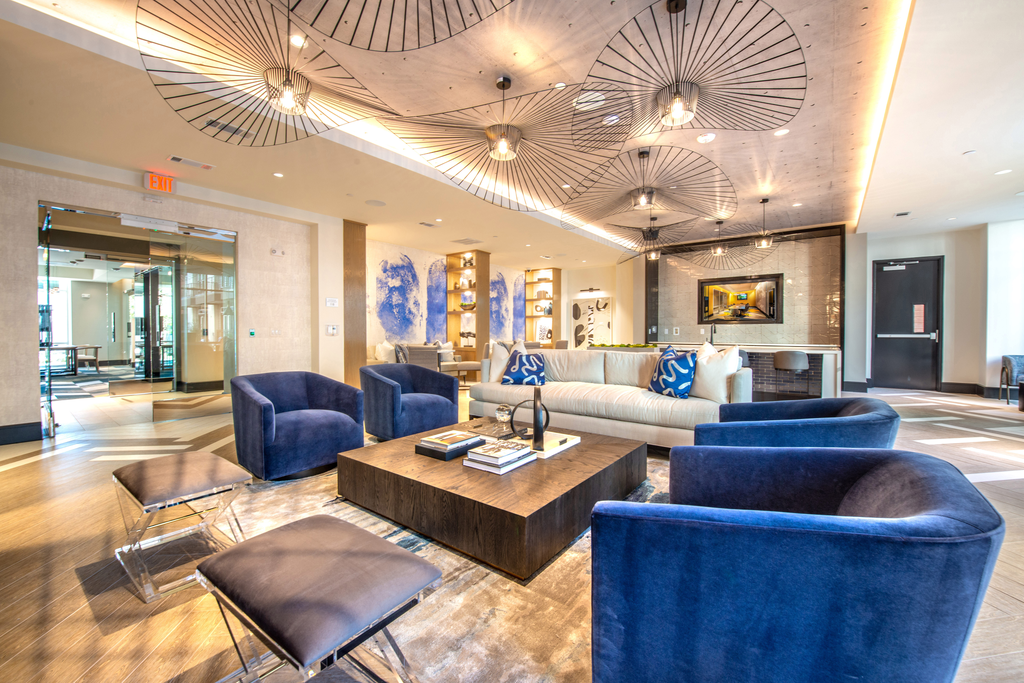 Clubhouse interior lounge with beige couch, blue chairs, brown seats, and wooden coffee table with accent rug and wood style flooring and unique ceiling hanging decor, wall mounted television