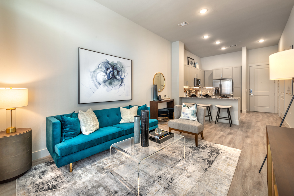 Staged livingroom with blue couch, accent rug, glass coffee table, cylindrical side table, gray chair and kitchen in background